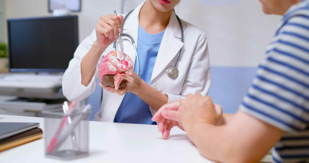 Closeup_of_young_female_doctor_with_white_coat_and_stethoscope_pen_pointing_and_explaining_conditions_related_to_brain_model_to_man_at_clinic.